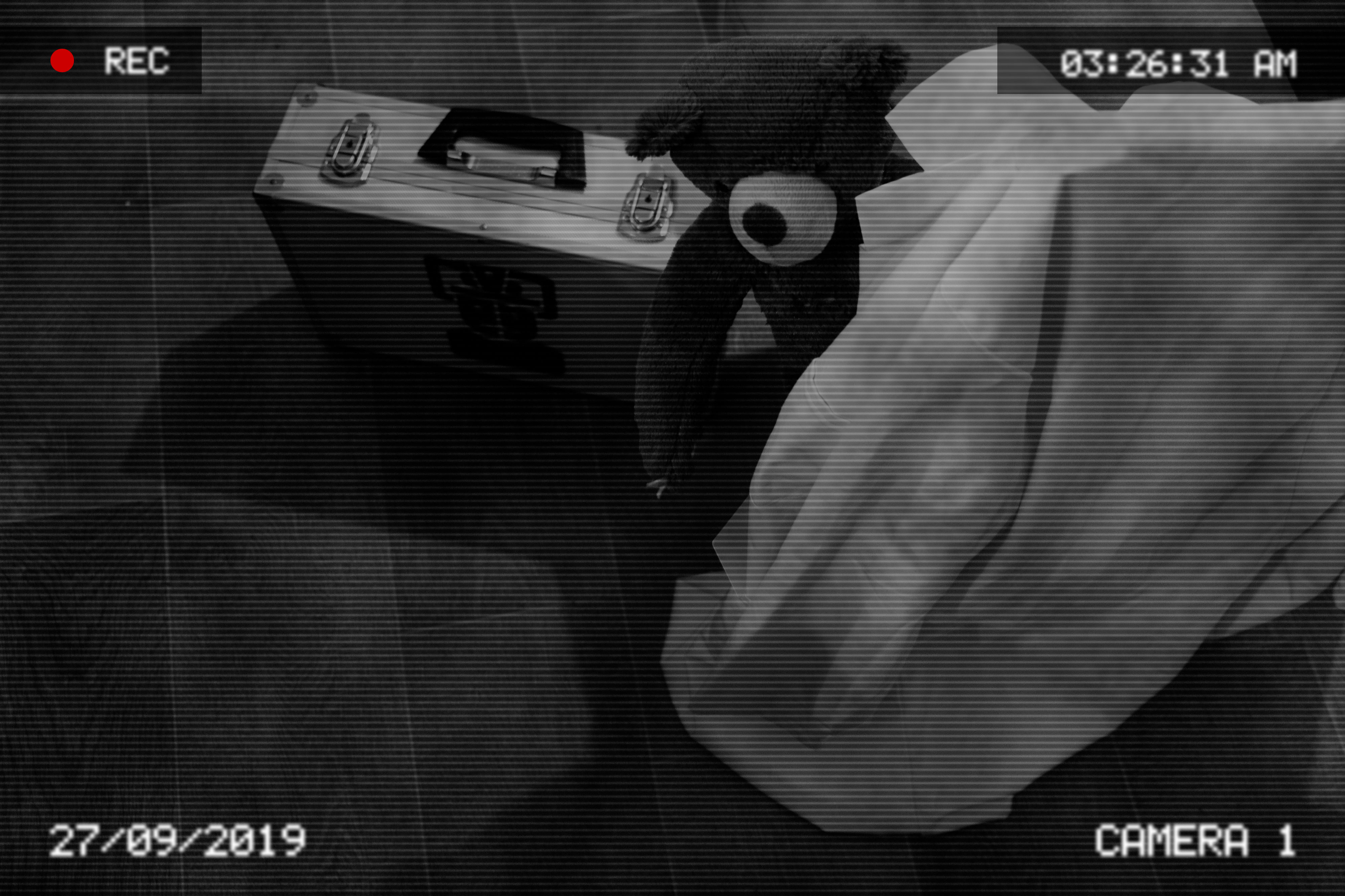 A teddy bear peaks out from behind cover near a silver case. The image appears to be from CCTV as it is interlaced and has a date stamp, a time stamp and the text "CAMERA !" embedded in it along with a record icon in the top left