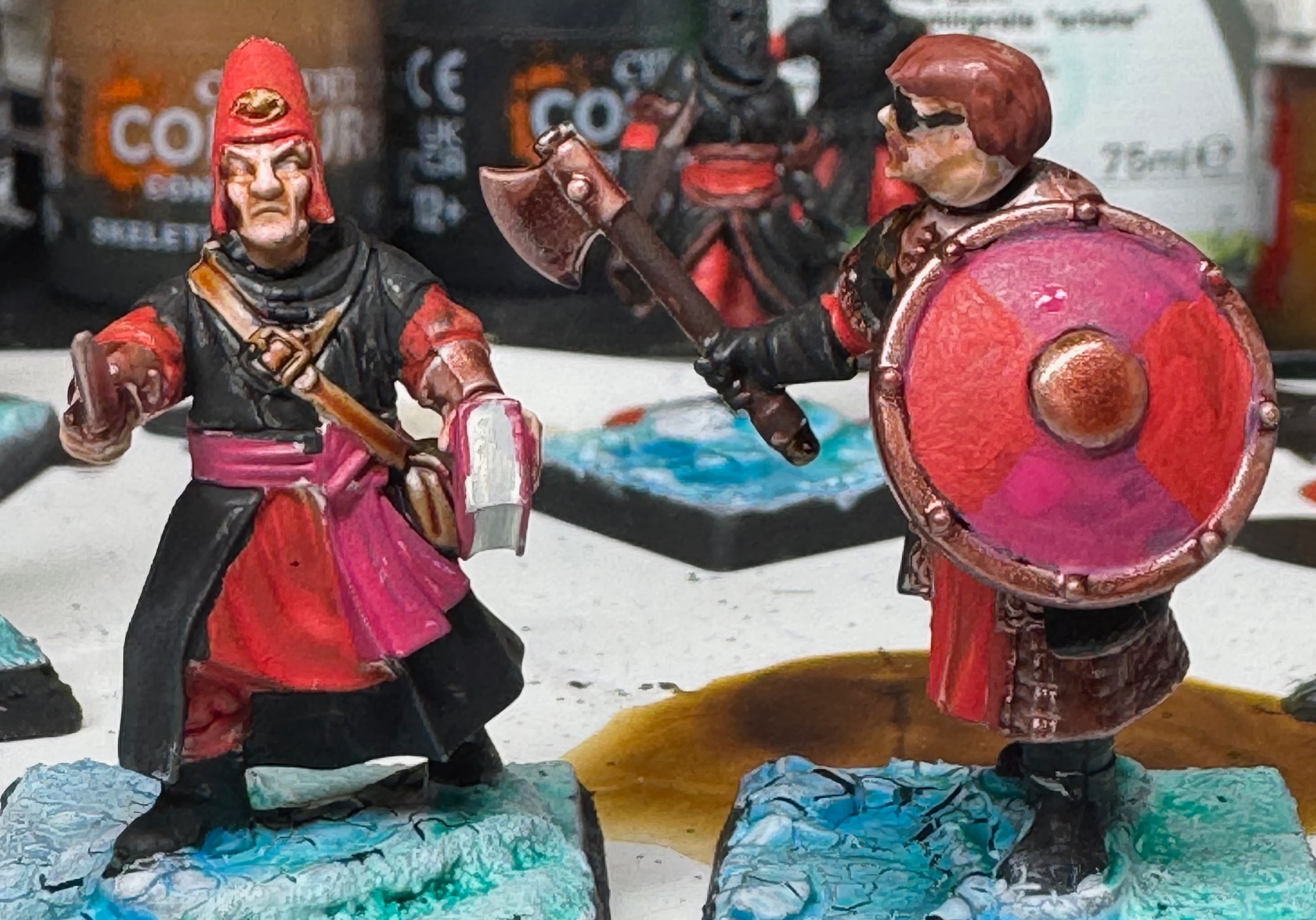 Two partially painted miniatures for Frostgrave; one is a very dodgy looking wizard, the other has an eyepatch and a round shield quartered into pink and red segments with a brass boss and rim.